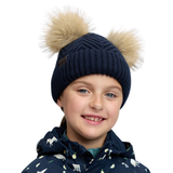 LeMieux AW26 Mini Double Pom Beanie Variant Navy. A smiling child wearing a navy ribbed knit beanie with two large beige faux-fur pom-poms and a navy hooded jacket with a white animal print, against a plain white background.