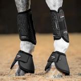 Horse legs wearing black protective leg wraps on a dirt ground.