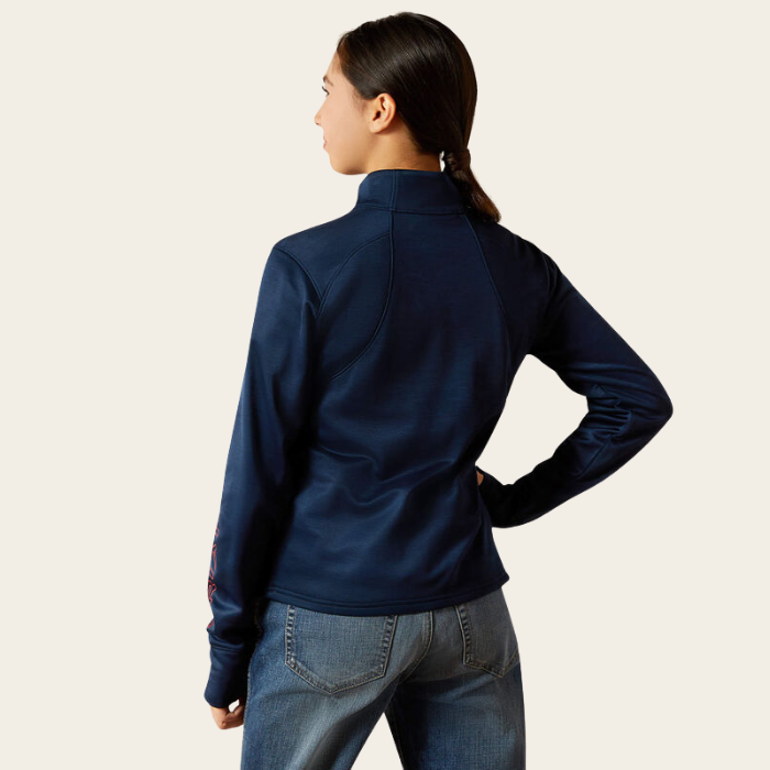 Ariat Youth Team 1/2 Zip Pullover Variant Navy. A woman shown from the back wearing a navy blue long-sleeve athletic quarter-zip jacket and blue jeans, set against a plain light beige studio background.