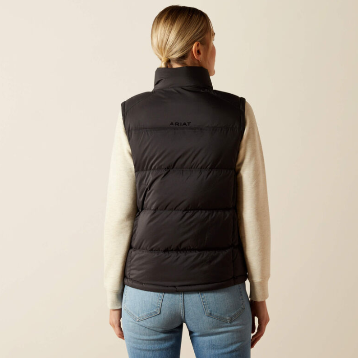 Ariat Women's Ultrapuff Down Vest Variant Black. A woman viewed from the back wearing a black puffer vest over a cream long-sleeve top and blue jeans, against a plain light beige studio background. The vest has a small ARIAT logo on the upper back.