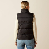 Ariat Women's Ultrapuff Down Vest Variant Black. A woman viewed from the back wearing a black puffer vest over a cream long-sleeve top and blue jeans, against a plain light beige studio background. The vest has a small ARIAT logo on the upper back.