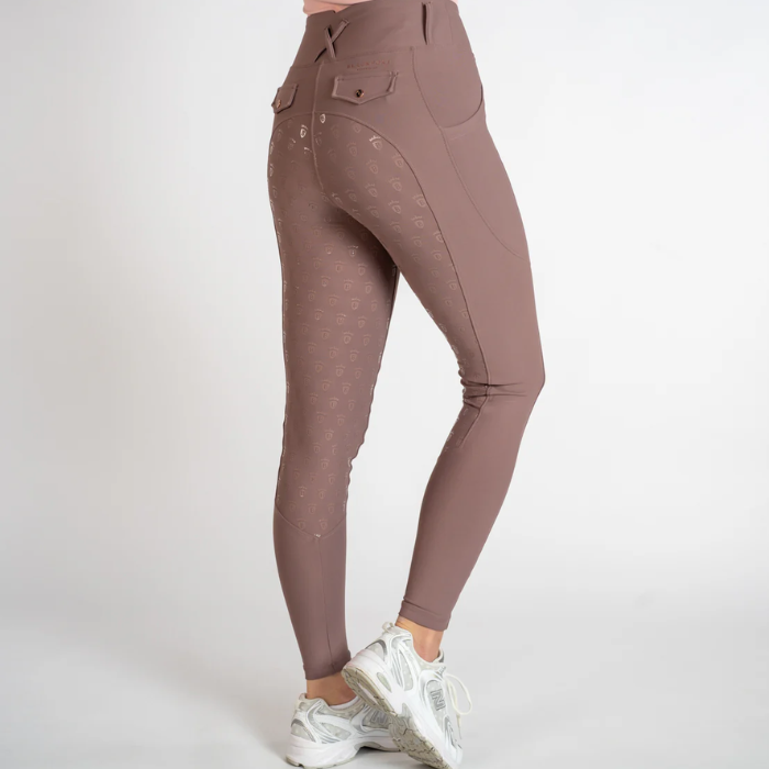 A person wearing high-waisted equestrian-style leggings in a dusty taupe-brown color, paired with white sneakers, shown from a side-rear angle against a light gray studio background.