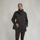 Holland Cooper Bramwell Waterproof Country Coat Variant Colour Bournville.
A woman wearing a black mid-length utility jacket with a high collar, paired with dark brown leggings and carrying a dark brown crossbody bag, standing against a textured light gray studio background.