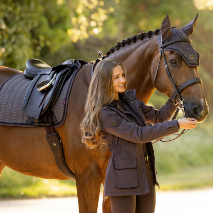 LeMieux AW26 Heritage Beatrice Waterproof Coat Variant Cocoa. A woman in a dark brown equestrian coat stands beside a brown horse outdoors, holding the horse's reins. The horse is outfitted with a dark brown bridle and matching horse blinders