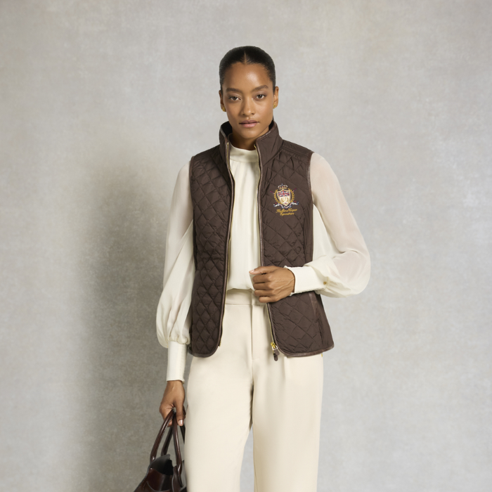 Holland Cooper Boughton Diamond Quilt Gilet Variant Colour Walnut.
A model stands against a textured gray studio backdrop wearing a dark brown quilted zip-front vest over a cream long-sleeve top and matching cream trousers. The vest has a small embroidered crest on the left chest.