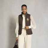 Holland Cooper Boughton Diamond Quilt Gilet Variant Colour Walnut.
A model stands against a textured gray studio backdrop wearing a dark brown quilted zip-front vest over a cream long-sleeve top and matching cream trousers. The vest has a small embroidered crest on the left chest.