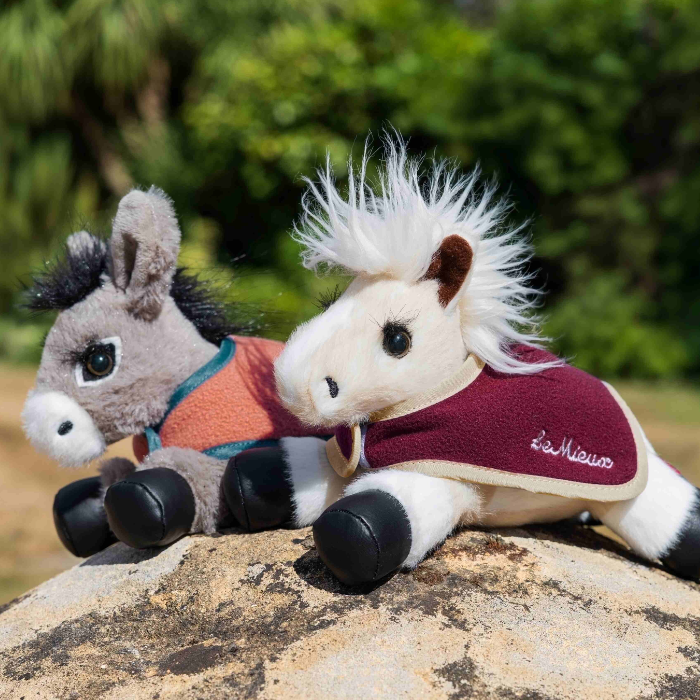 Two plush stuffed animals, a gray horse and a white-and-black horse, posed together on a rock outdoors with a blurred green natural background. The horse wears a burgundy fleece saddle blanket with white trim and the script brand name 'LeMieux'.