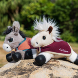 Two plush stuffed animals, a gray horse and a white-and-black horse, posed together on a rock outdoors with a blurred green natural background. The horse wears a burgundy fleece saddle blanket with white trim and the script brand name 'LeMieux'.