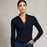 Holland Cooper Zoe Quarter Zip Knit Variant Colour Ink Navy.
A woman modeling a dark navy ribbed knit half-zip sweater with a beige lining and gold-tone zipper pull, paired with blue jeans against a textured gray studio background.