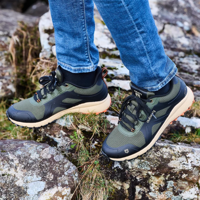 A person wearing blue jeans and a black top stands outdoors on rocky ground, showing a pair of green and black trail shoes with beige midsoles and orange outsoles. The brand name 'DUBARRY' is visible on the side of the shoe.