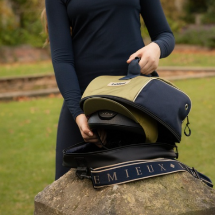 LeMieux AW26 Hat Bag Varient Gooseberry. A person outdoors holds an olive green and navy blue helmet bag open over a stone, with a black helmet inside and a branded shoulder strap visible. The background shows grass and trees.
