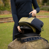 LeMieux AW26 Hat Bag Varient Gooseberry. A person outdoors holds an olive green and navy blue helmet bag open over a stone, with a black helmet inside and a branded shoulder strap visible. The background shows grass and trees.