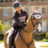 LeMieux AW26 Heritage Fly Hood Variant Wild Rose. A woman in equestrian attire rides a chestnut horse outdoors in front of a brick building. The rider wears a dark helmet and a dark charcoal equestrian jacket with a pinkish-beige panel and the brand name 'LeMieux' visible.