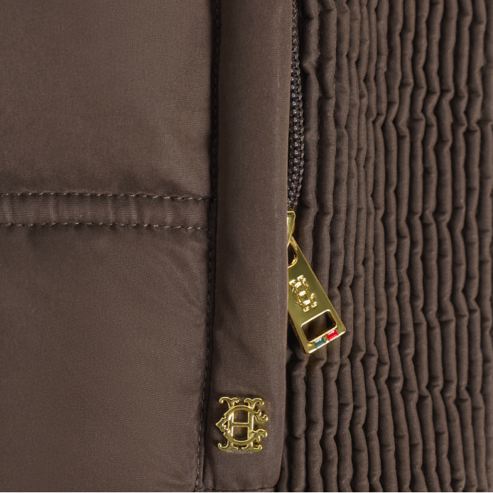 Holland Cooper Cranborne Training Gilet Variant Chocolate. Close-up detail of a brown quilted jacket showing a gold-tone zipper pull and a matching gold-tone crest logo charm.