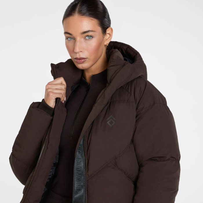 Aztec Diamond Matt Puffer AW26 Variant Espresso. A woman wearing a dark brown puffer jacket with a high collar and a subtle geometric logo on the chest, photographed against a clean light gray studio background.