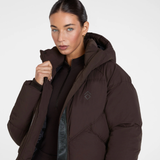 Aztec Diamond Matt Puffer AW26 Variant Espresso. A woman wearing a dark brown puffer jacket with a high collar and a subtle geometric logo on the chest, photographed against a clean light gray studio background.