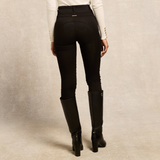 Holland Cooper Jodhpur Jean Variant Black. Rear view of a model wearing black riding breeches with gold button details on the back pockets, paired with a white long-sleeve top and black knee-high boots, against a textured beige studio backdrop.