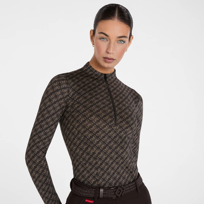 A woman modeling a fitted long-sleeve quarter-zip top with an allover repeating monogram pattern in dark brown and beige, paired with a black belt against a clean light gray studio background.