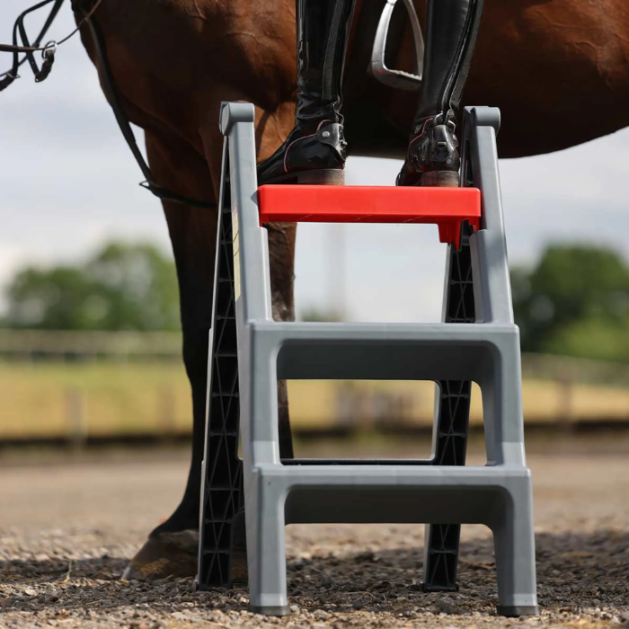 A gray plastic horse step ladder with a bright red second step, shown outdoors with a horse and rider's legs visible.