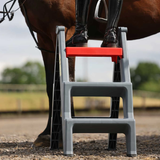 A gray plastic horse step ladder with a bright red second step, shown outdoors with a horse and rider's legs visible.