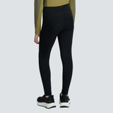 Rear view of a model wearing black athletic leggings with textured paneling and a small visible brand logo near the waistband, paired with black sneakers against a light gray studio background.
