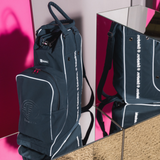 A dark navy blue boot/ hat bag with white piping and a gray circular logo on the front pocket, featuring dual top handles and shoulder straps, shown on a mirrored shelf. 