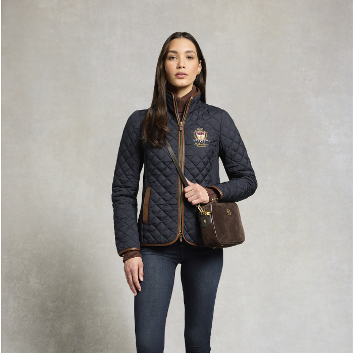 Holland Cooper Boughton Diamond Quilt Jacket Variant Colour Ink Navy.
A woman modeling a dark navy quilted zip-up jacket with brown trim and a small embroidered crest on the left chest, paired with dark jeans and a brown crossbody bag against a textured gray studio background.