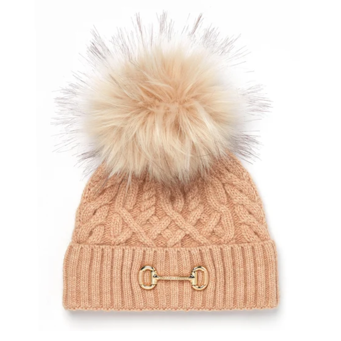 Holland Cooper Horsebit Bobble Hat Variant Camel. A cable-knit beanie in a warm peach-beige color, featuring a large fluffy faux-fur pom-pom in a matching light beige tone and a gold-tone horsebit detail on the front cuff.