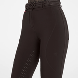 Close-up of a person wearing brown equestrian riding breeches with textured grip panels on the lower legs and a matching black patterned riding belt, shown against a light gray studio background.