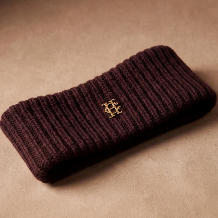 A dark burgundy ribbed knit headband with a small gold interlocking monogram logo, displayed on a textured beige surface.