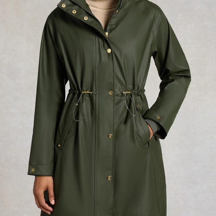 Holland Cooper Chartwell Rain Parka Variant Hunter Green. An olive green hooded raincoat with gold-tone snap buttons and drawstring details, worn by a model against a textured gray background.