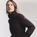 Close up of a lady wearing a brown jacket with a HC logo on the sleeve against a grey background. 