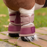 LeMieux AW26 Toy Pony Over Reach Boots Variant Colour Blackberry. A close up of a toy pony wearing blackberry coloured leg wraps on faded garden background
