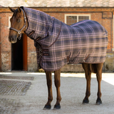 A dark horse standing on a yard, wearing a quilted horse blanket with a blue, gray, and pink plaid pattern and a matching neck cover.