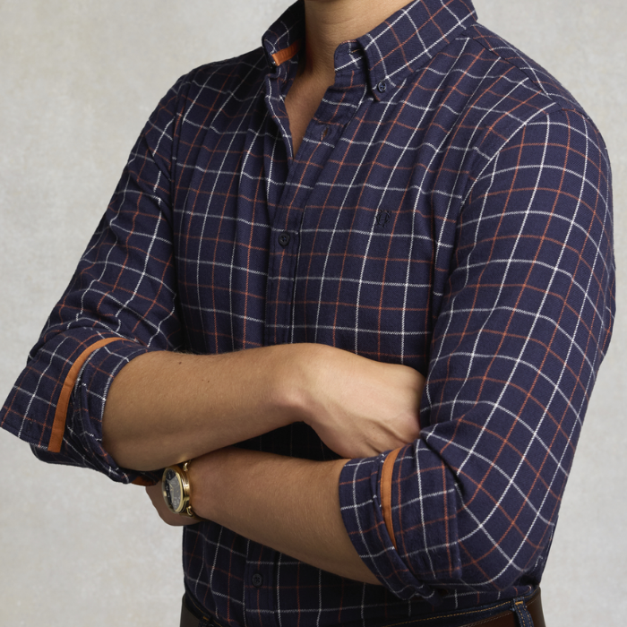 Holland Cooper Men's Classic Fit Brushed Cotton Shirt Variant Copper Navy Check. A man wearing a navy blue long-sleeve plaid button-down shirt with white and orange grid lines, shown from the neck down with arms crossed.