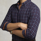 Holland Cooper Men's Classic Fit Brushed Cotton Shirt Variant Copper Navy Check. A man wearing a navy blue long-sleeve plaid button-down shirt with white and orange grid lines, shown from the neck down with arms crossed.