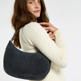 A woman wearing a cream cable-knit sweater carries a dark navy blue suede crescent shoulder bag with gold-tone hardware against a plain white background.