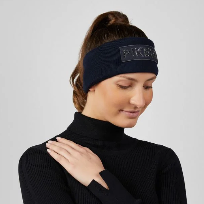 Pikeur AW26 Cotton Headband Variant NightBlue. A woman wearing a dark navy knit headband with a rectangular logo patch that reads 'PIKEUR' in raised letters, paired with a black ribbed turtleneck sweater against a light gray studio background.
