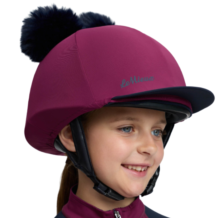 LeMieux AW26 Mini Double Pom Hat Silk Variant Colour Blackberry.
A child wearing a burgundy riding helmet with a black pom-pom and visor, featuring the 'La Mignonne' brand name embroidered on the side, set against a plain white background.