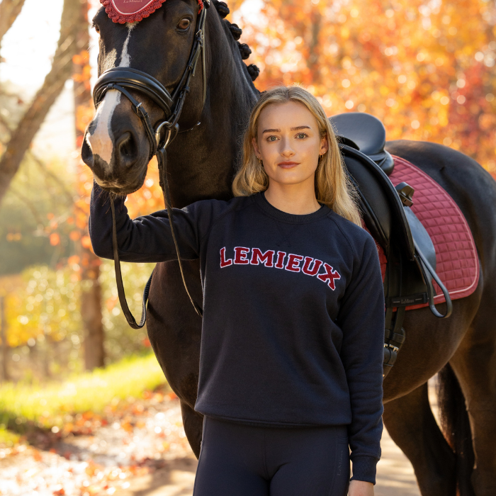 LeMieux AW26 Vida Varsity Crew Neck Sweat Variant Colour Navy.
A woman stands outdoors beside a dark horse wearing a bridle and saddle with a pink quilted pad. The woman is wearing a navy sweatshirt with 'LEMIEUX' printed across the chest in red and white, paired with navy leggings.