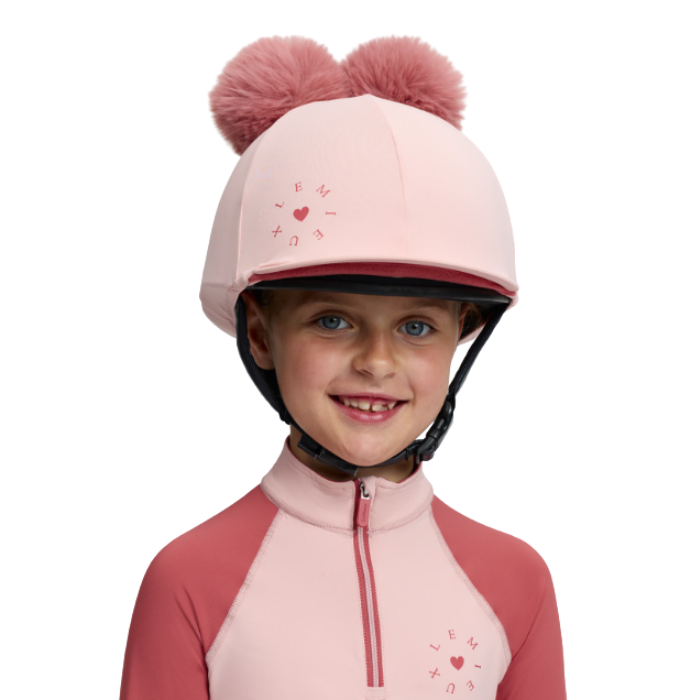 LeMieux AW26 Mini Double Pom Hat Silk Variant Colour Petal.
A young child wearing a light pink riding helmet with two pink pompoms and a matching pink and coral long-sleeve riding jersey, photographed against a clean white background.