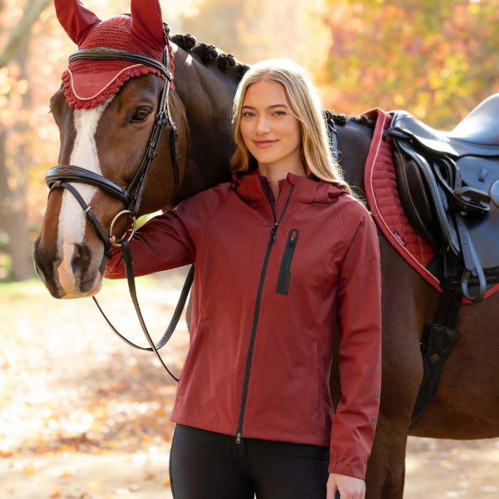 LeMieux AW26 Athena Waterproof Lightweight Jacket Varient Russet. A woman stands beside a brown horse outdoors, wearing a fitted zip-front jacket in a muted brick red color with black trim. The horse has a red blind and a black saddle, and the background shows a softly blurred autumn landscape with fallen leaves.