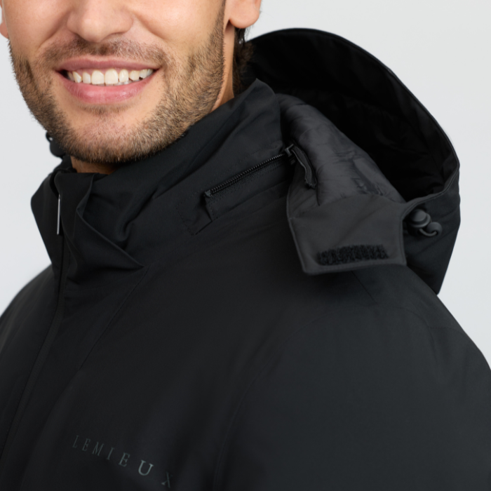 LeMieux AW26 Mens Waterproof Training Coat Varient Black. Close-up of a man wearing a black hooded jacket, showing the collar, zipper, hood strap, and part of the chest area with subtle branding text.