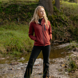 LeMieux AW26 Athena Waterproof Lightweight Jacket Varient Russet. A woman stands outdoors near a small stream in a wooded area, wearing a fitted zip-front jacket in a muted brick red or burgundy color with black pants.