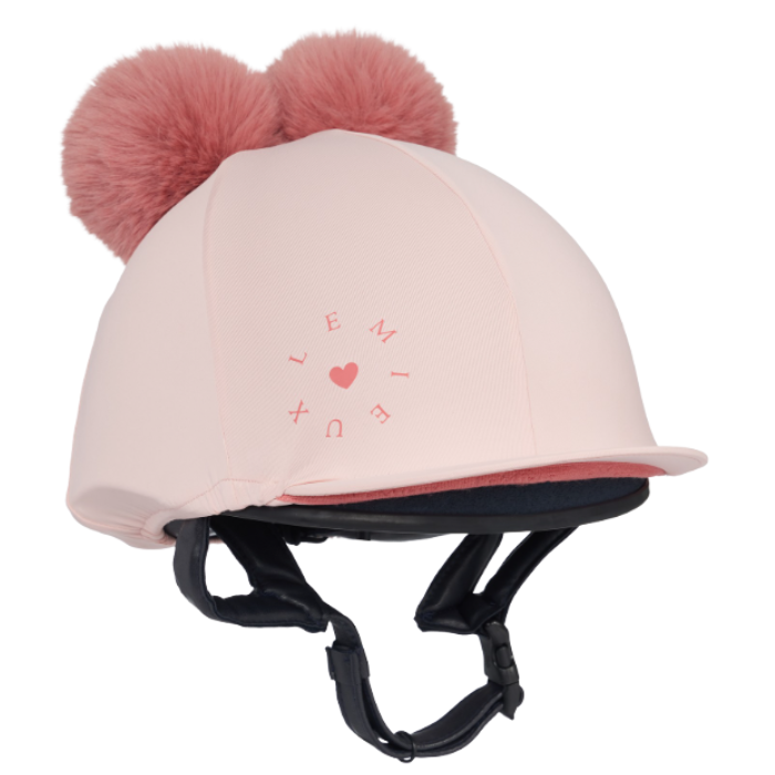 LeMieux AW26 Mini Double Pom Hat Silk Variant Colour Petal.
A light pink equestrian riding helmet cover with two fluffy pink pom-poms on top, featuring a pink heart and letter design on the front, and black chin straps, shown against a plain white background.