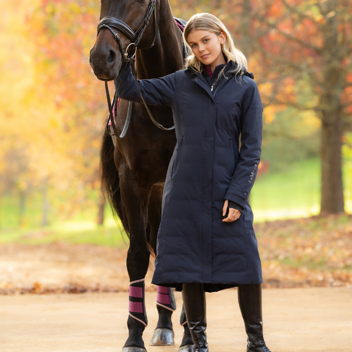 LeMieux AW26 Erin Waterproof Padded Coat Varient Navy. A woman wearing a long navy equestrian coat stands beside a dark horse in an outdoor park setting with autumn trees in the background.