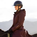 LeMieux AW26 Erin Waterproof Padded Coat Varient Bramble. A woman wearing a dark navy equestrian helmet and a maroon hooded riding jacket with 'Le Mieux' embroidered on the sleeve, riding a horse against a soft, hazy outdoor background.