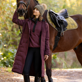 LeMieux AW26 Erin Waterproof Padded Coat Varient Bramble. A woman in a long burgundy quilted riding coat, black riding breeches, and a beige knit beanie stands beside a brown horse outdoors in a wooded setting.