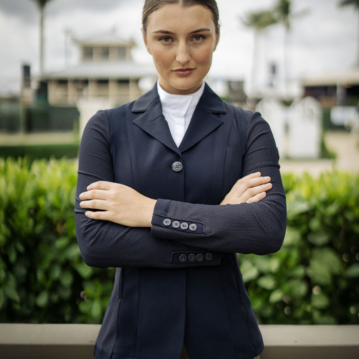 Woman in equestrian attire standing with arms crossed outdoors.