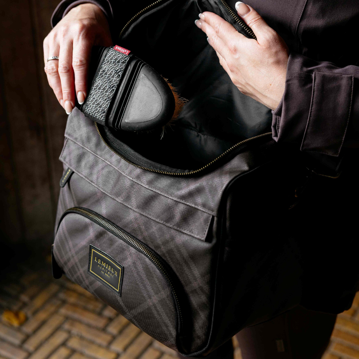 A person holds open a dark gray plaid grooming bag, showing its interior and a small black grooming brush inside. The bag has a front zippered pocket with a gold-toned 'LeMieux Heritage' logo patch.